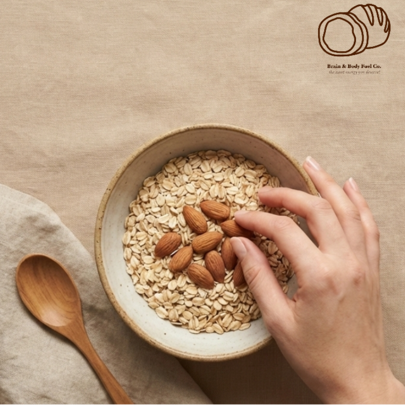 It starts the story of Brain and body fuel .co. Bowl of oats with almonds on a beige surface, hand reaching in, wooden spoon beside, brand logo at top.